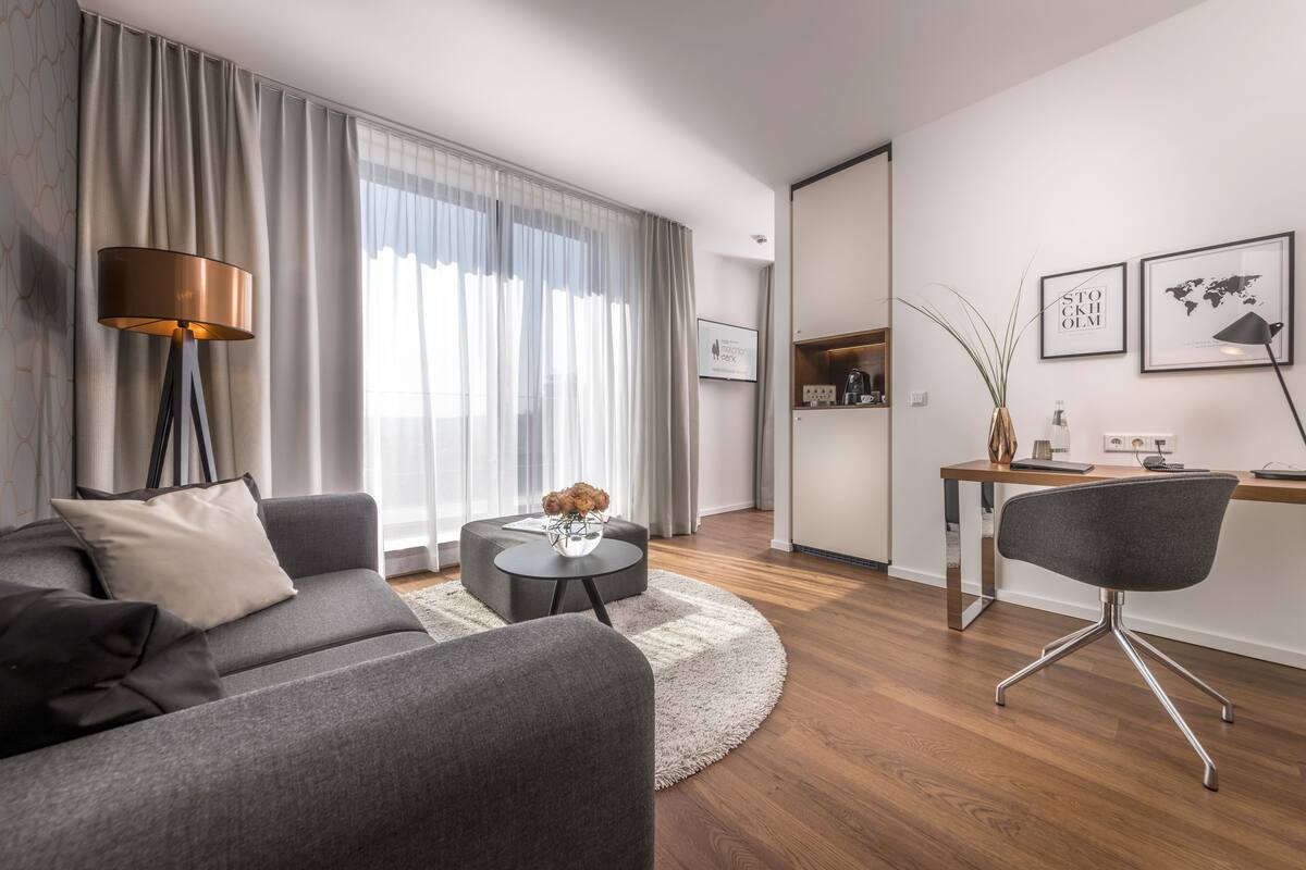 A modern living room with a grey sofa, a round coffee table, and a grey armchair. There is a lamp, a small table with decorative items, and a view of the kitchen through a glass door.