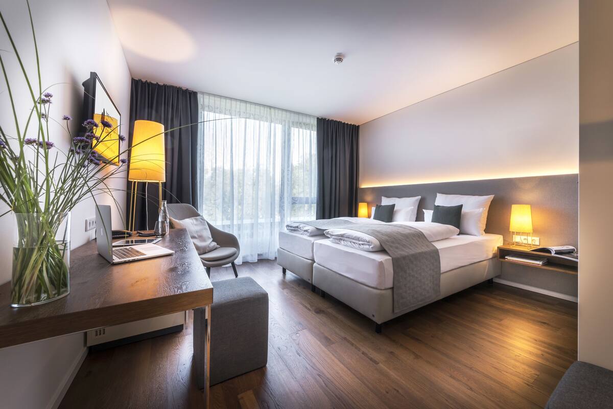 A modern hotel room with a wooden floor, a desk with a laptop, a comfortable bed with pillows, a bedside table with a lamp, and a large window with curtains.