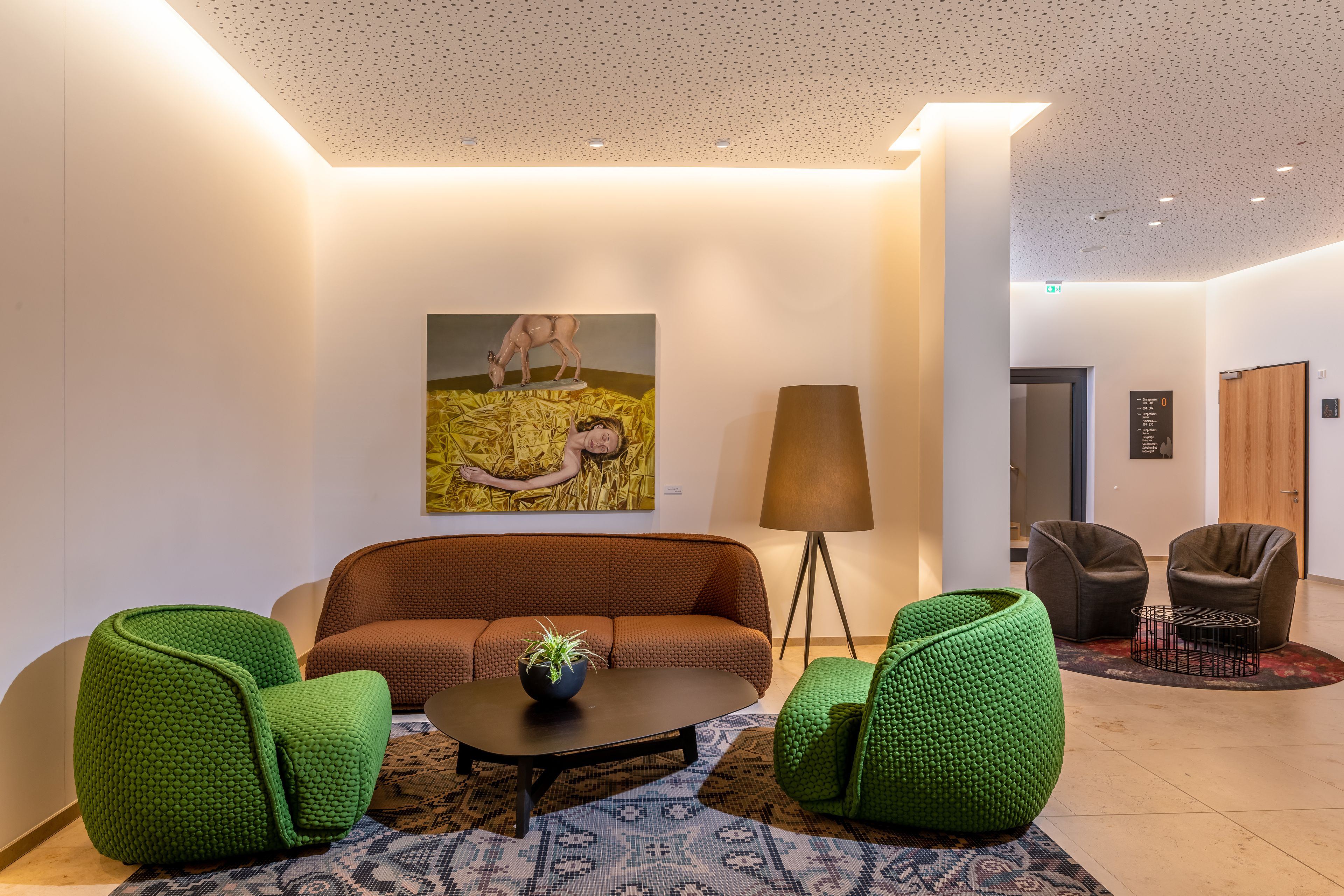 A modern lobby with a brown sofa, green pouf chairs, a black coffee table, and a large painting on the wall.