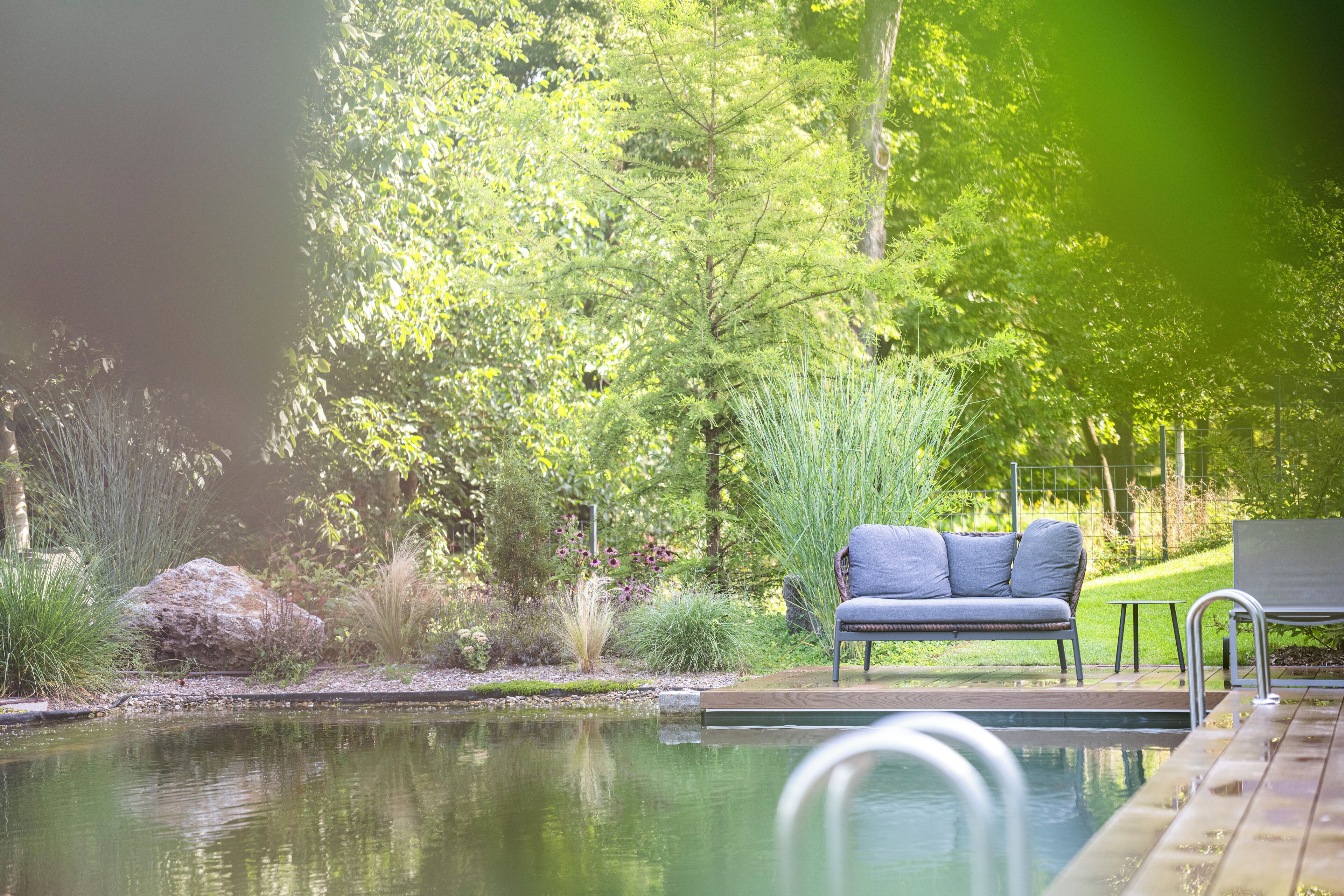 A pool with a wooden deck, a grey sofa, and lush greenery in the background.