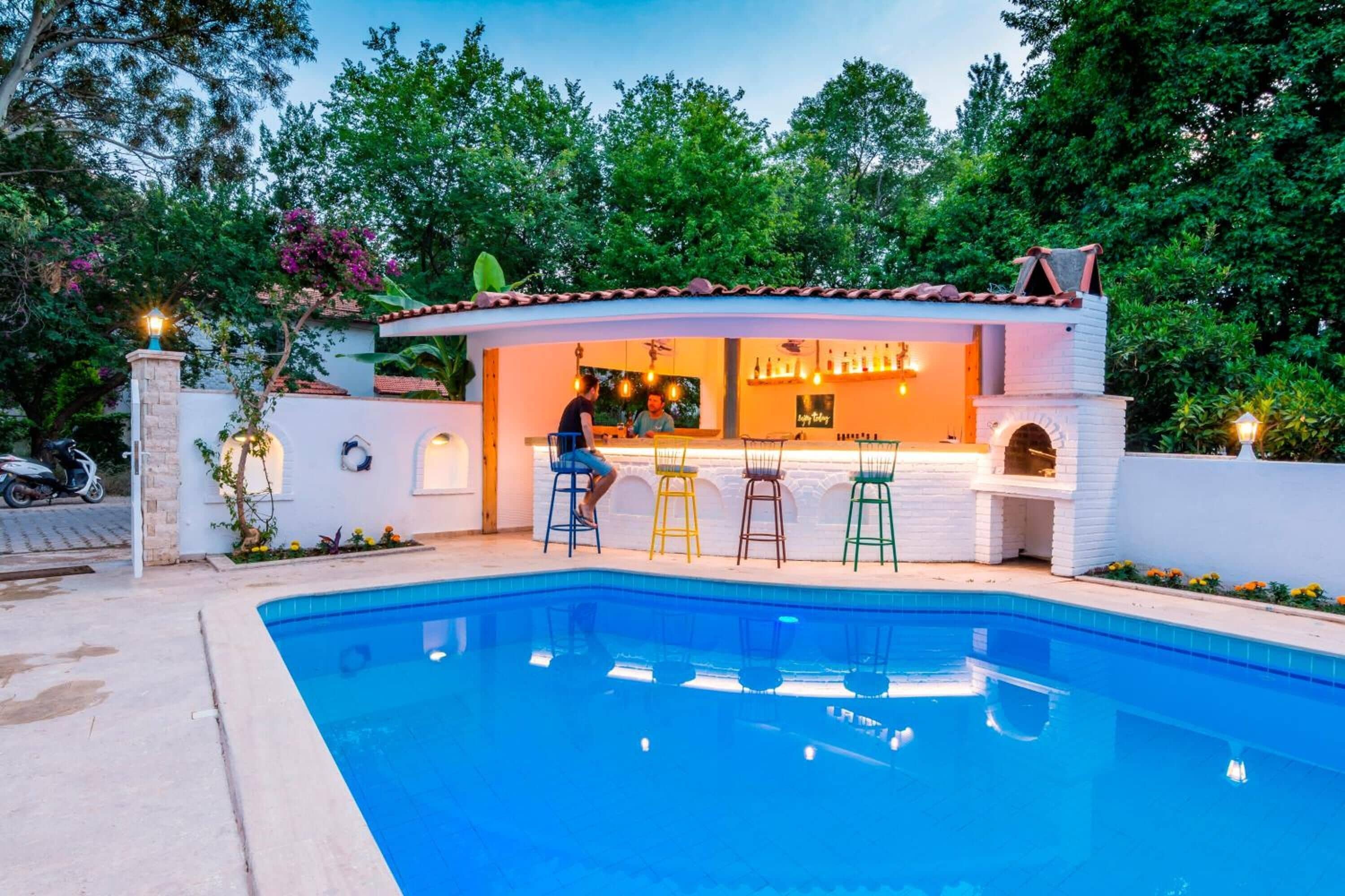 Un bar junto a la piscina con fachada blanca, varios taburetes y una persona sentada.
