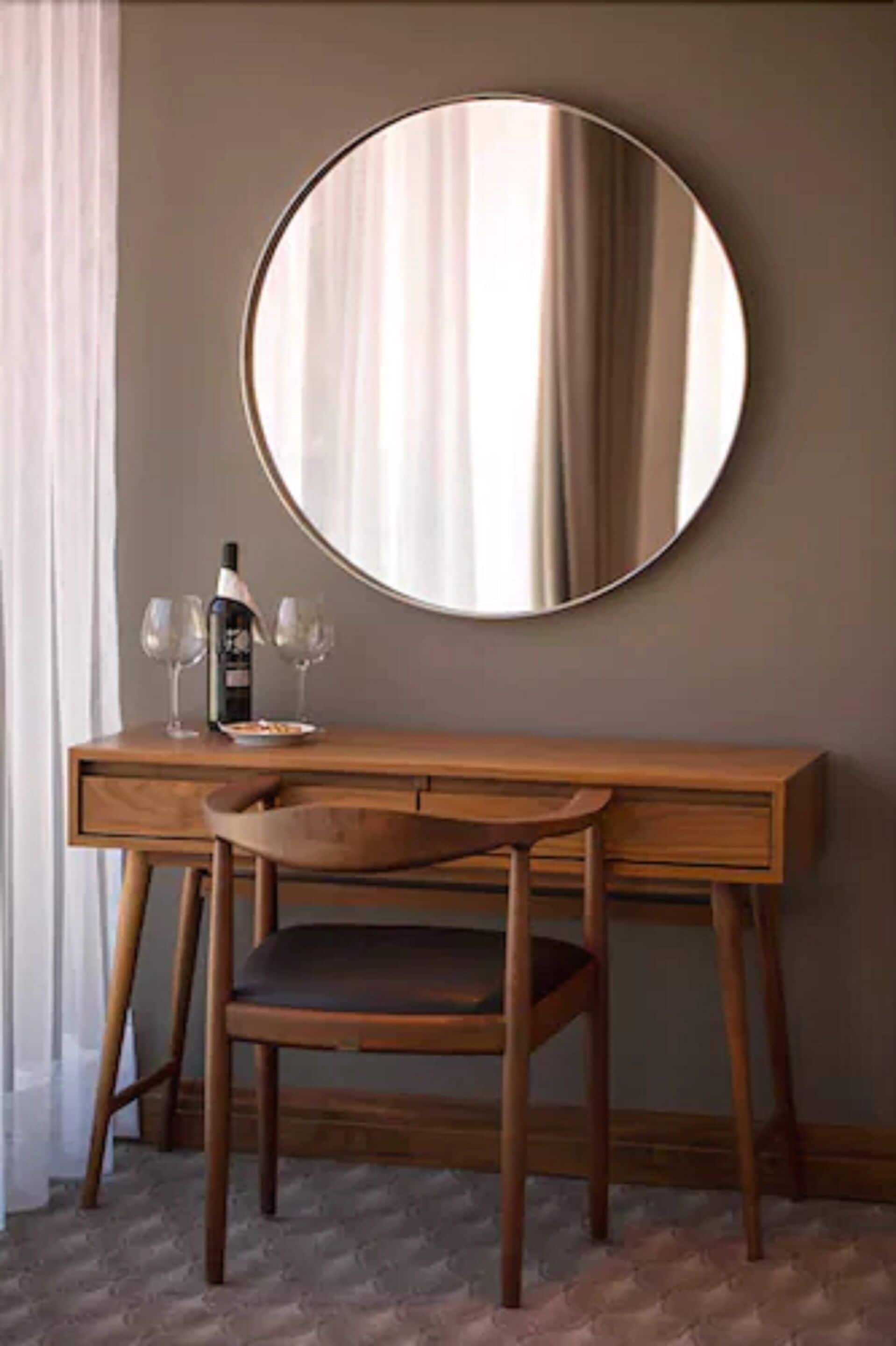 A wooden desk with a round mirror, two wine glasses, and a bottle of wine.