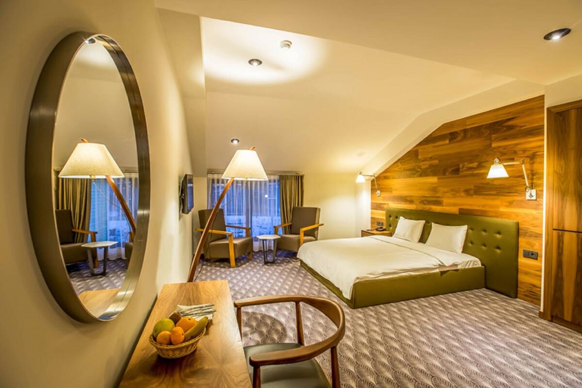 A hotel room with a large bed, a desk with a lamp, a chair, a mirror, and a view of the city through the window.