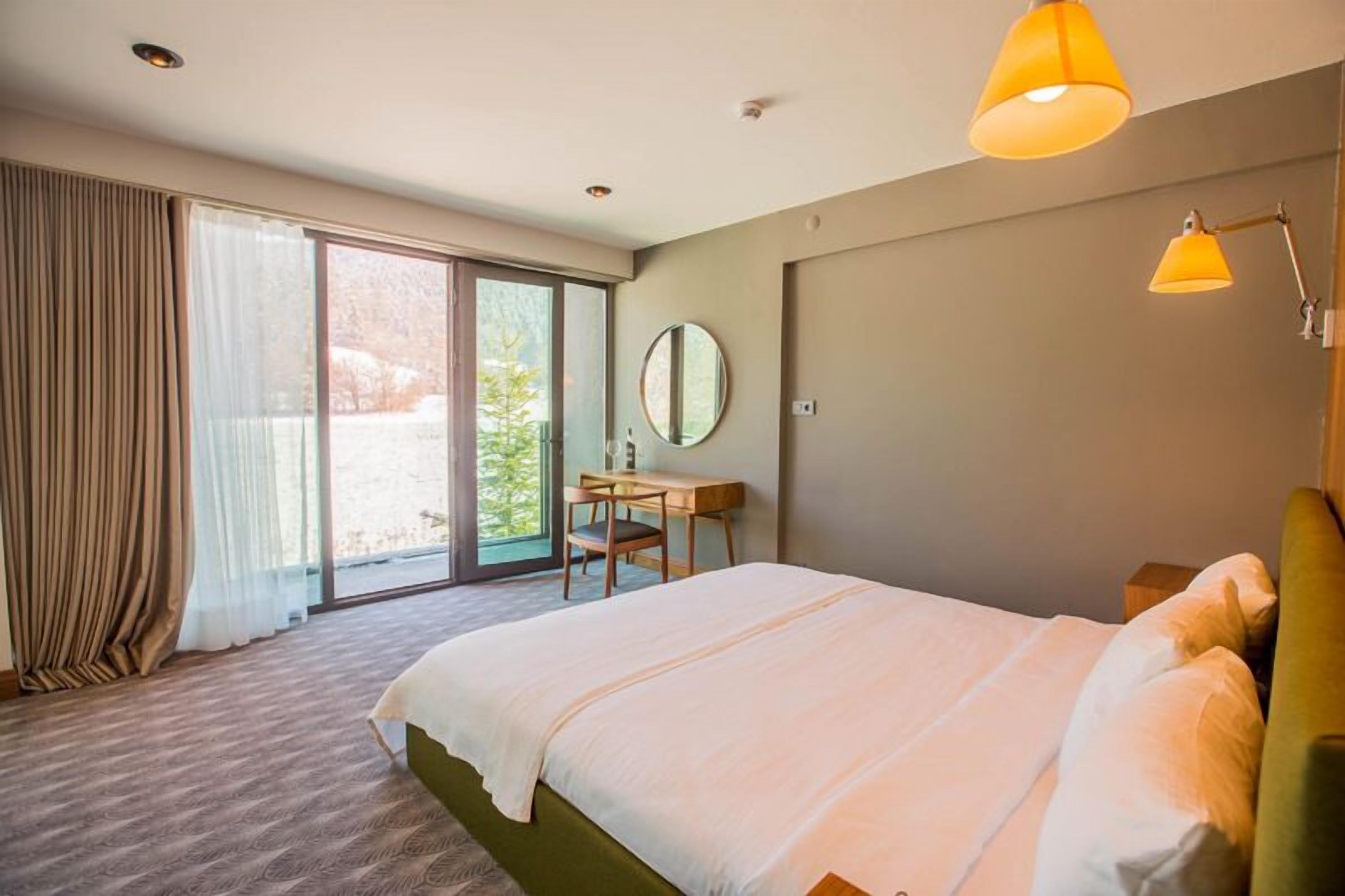 Quarto de hotel com uma cama grande, uma escrivaninha, uma cadeira e vista através de uma porta de vidro deslizante.