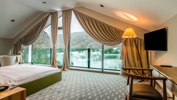 A hotel room with a large bed, a desk, a chair, and a TV. The room has large windows with curtains and a view of greenery outside. - Abant Lotus Otel (Bolu)