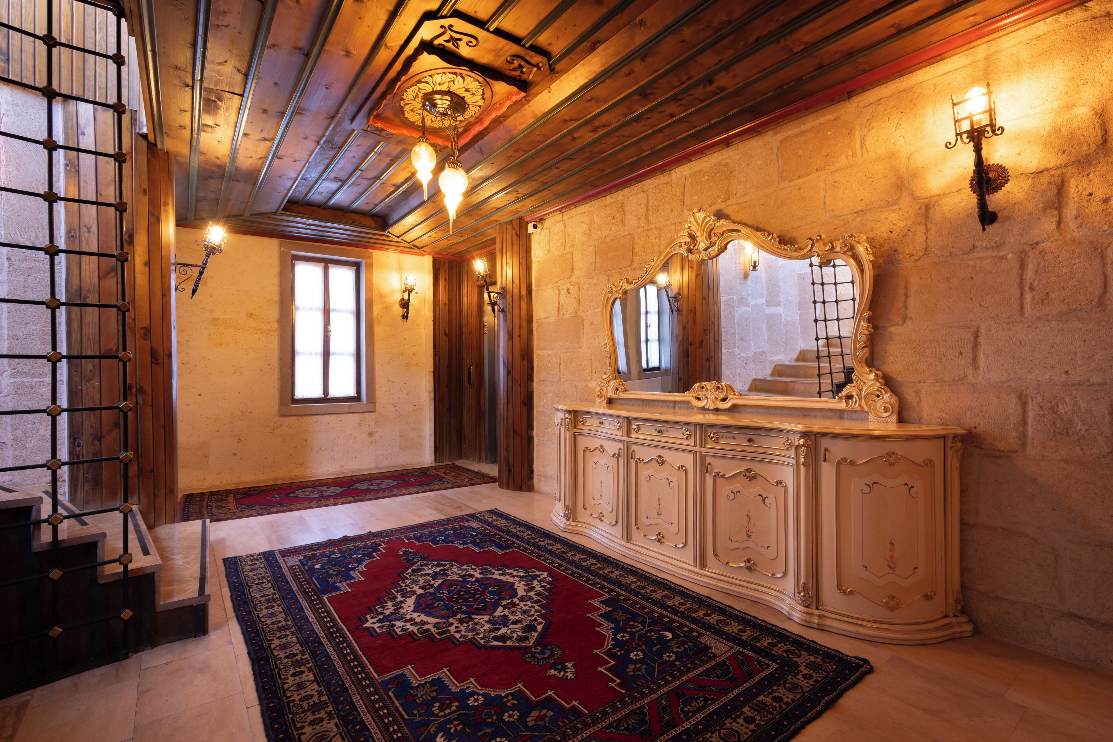 A room with a wooden ceiling, a large ornate mirror, a patterned rug, and a light fixture on the wall.