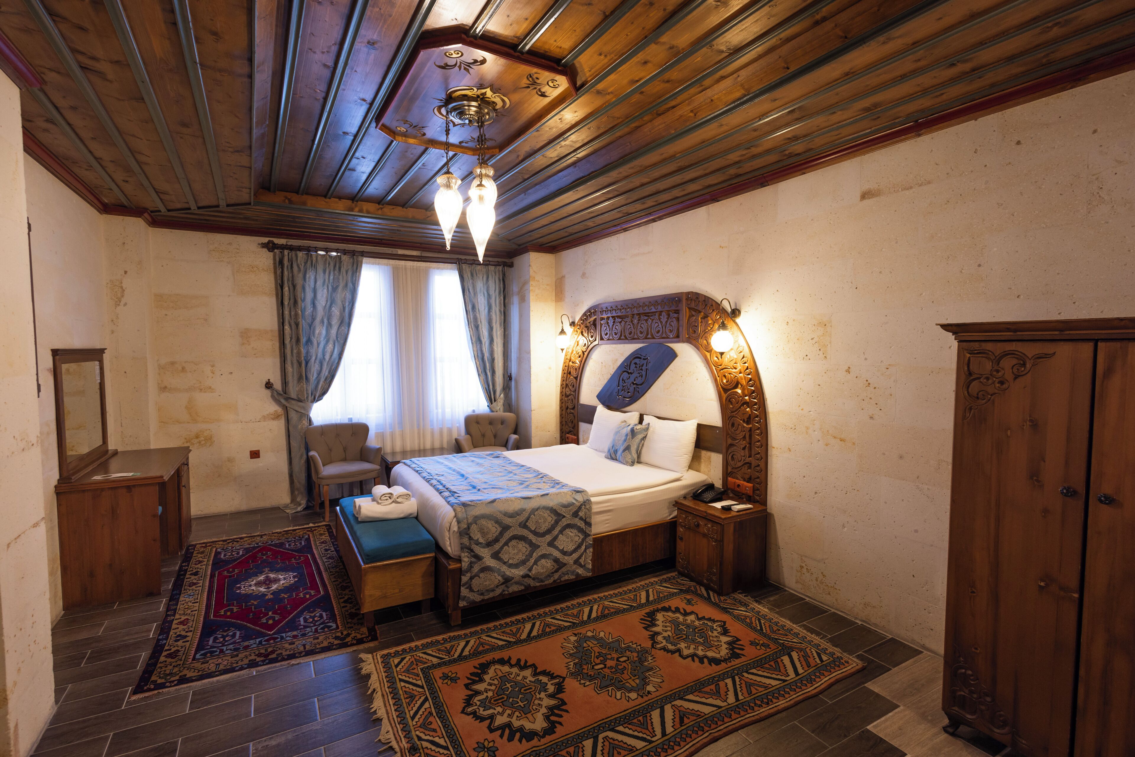 A room with a bed, a nightstand, a wooden wardrobe, a chair, a desk, a mirror, and a patterned rug.