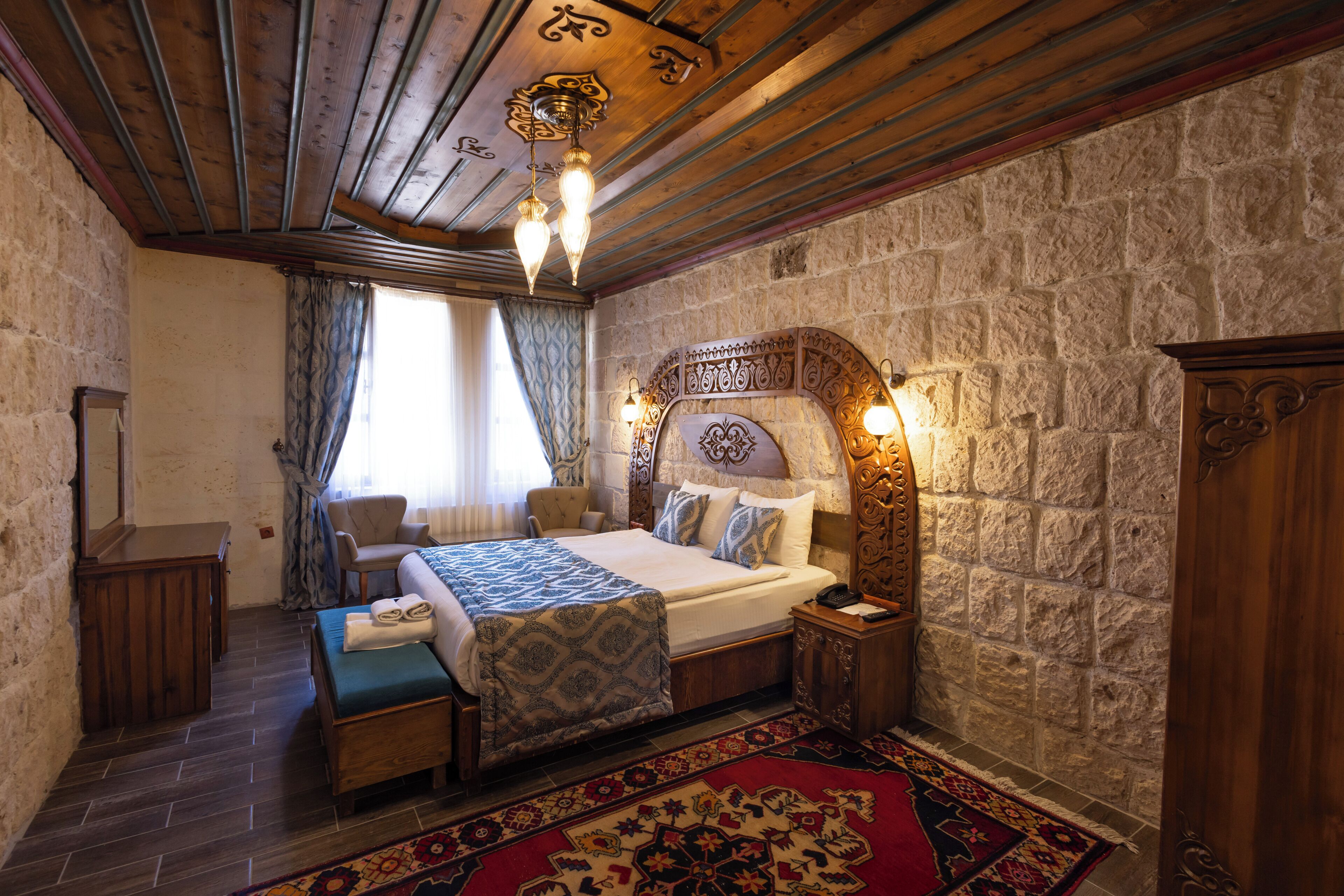 Un dormitorio con pared de piedra, vigas de madera en el techo, una cama con ropa de cama estampada, una mesita de noche y una silla.