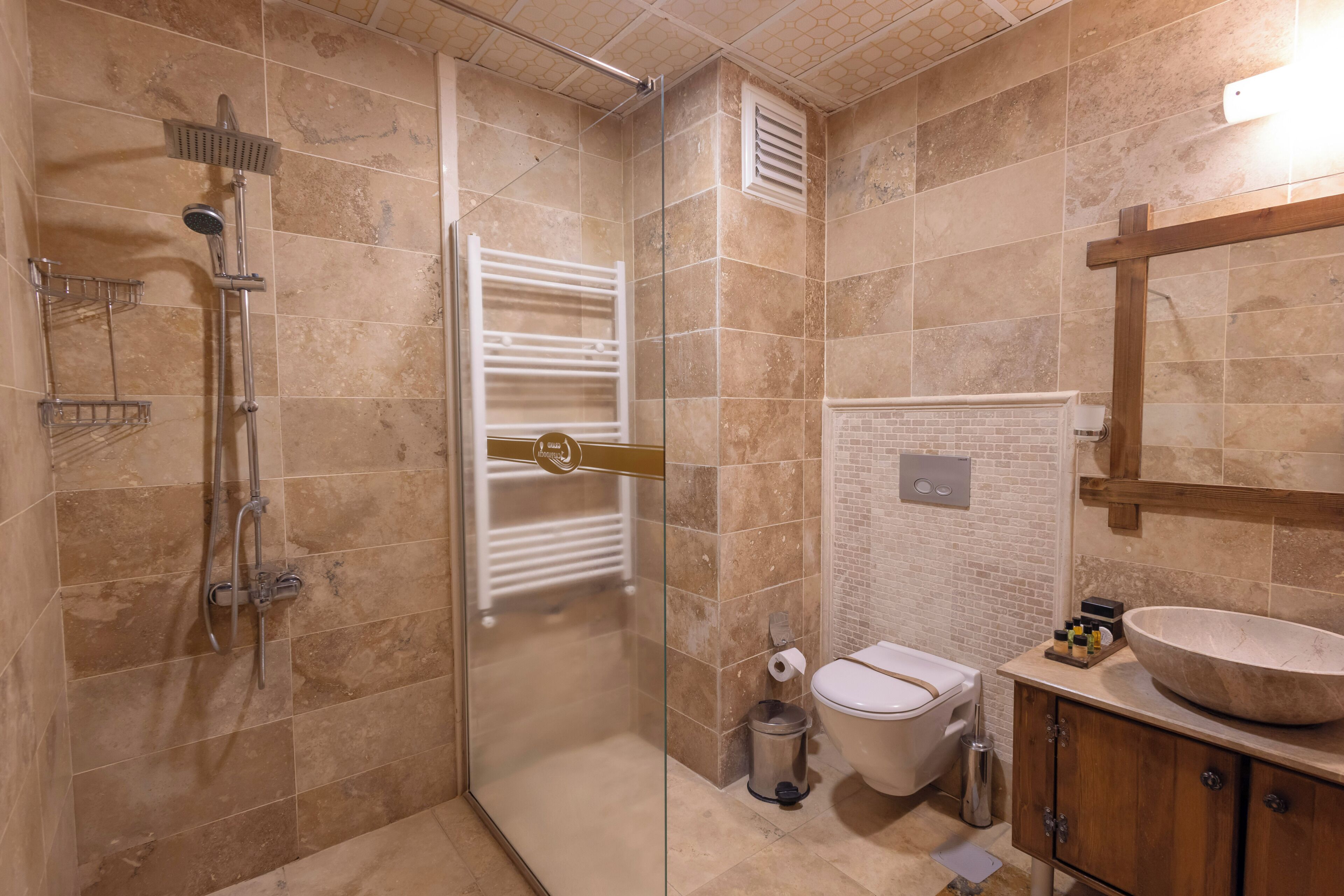 Un baño con ducha de vidrio, inodoro blanco, tocador de madera con un lavamanos grande y un perchero para toallas.