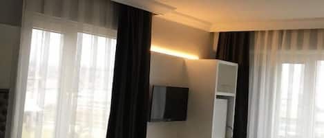 A hotel room with a bed, a desk with a chair, a TV mounted on the wall, and a window with curtains.