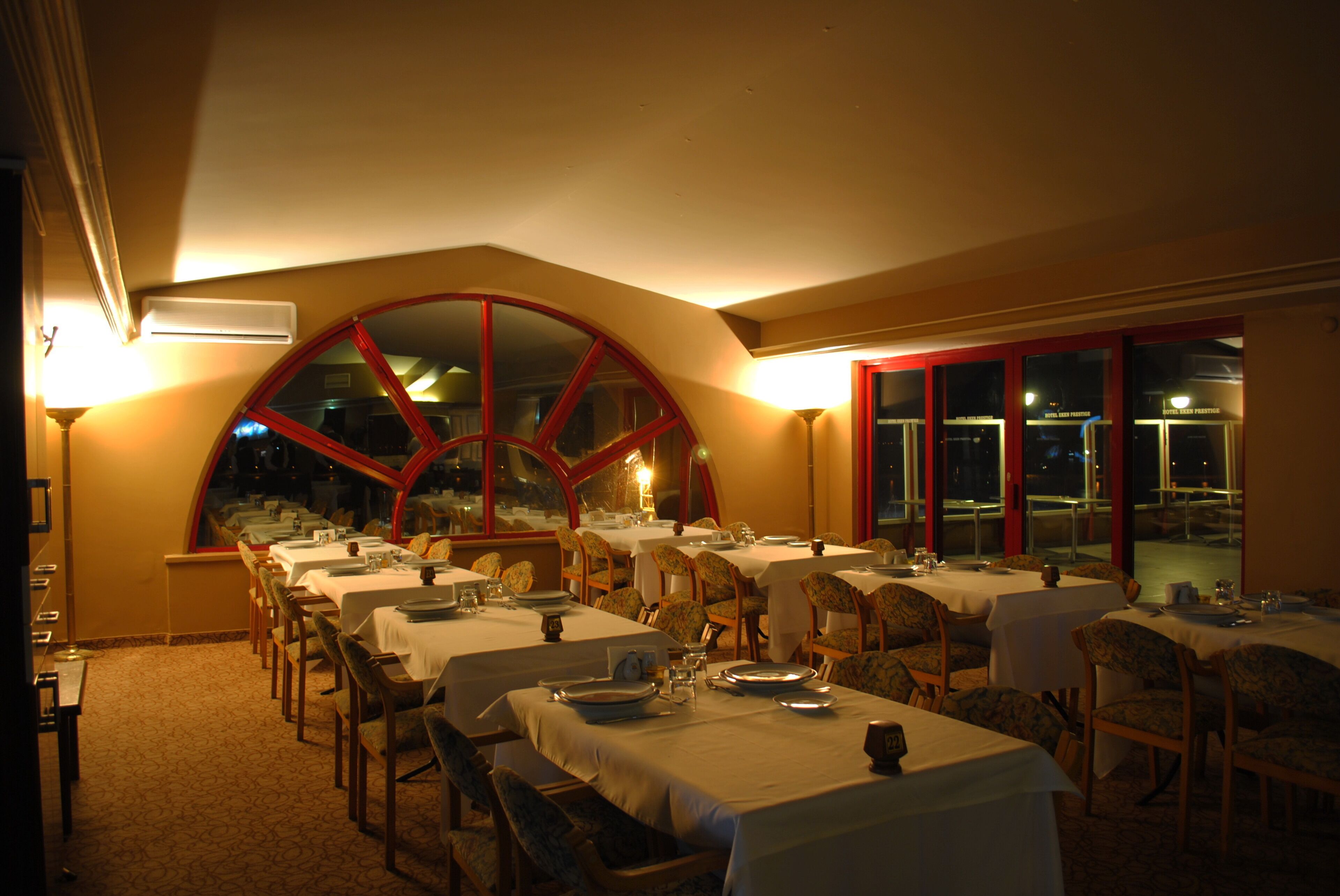 Un restaurante con varias mesas dispuestas para comer, amplias ventanas con marcos rojos e iluminación interior cálida.
