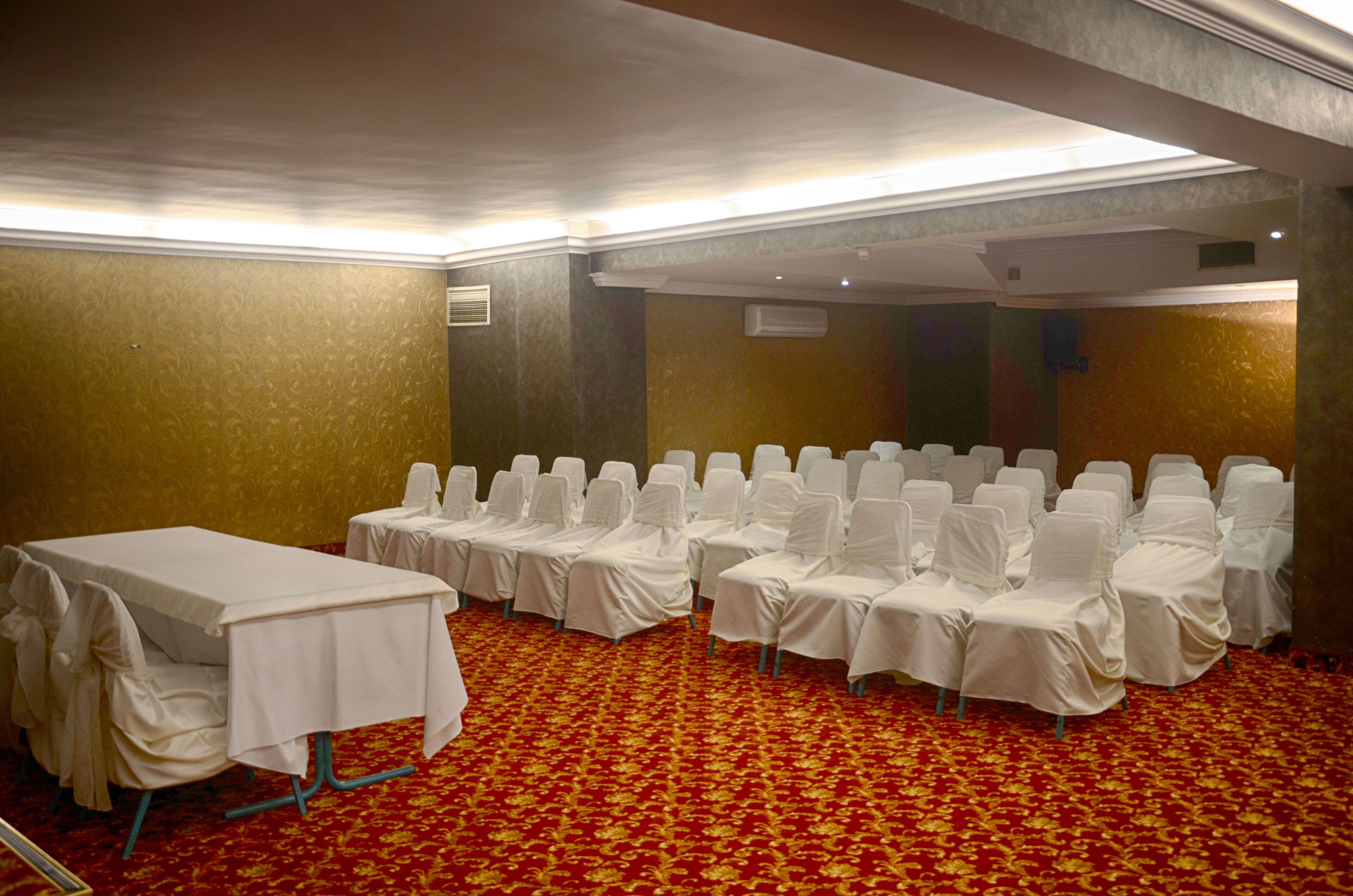 A conference room with rows of white chairs and a table, carpeted in red with a gold pattern.