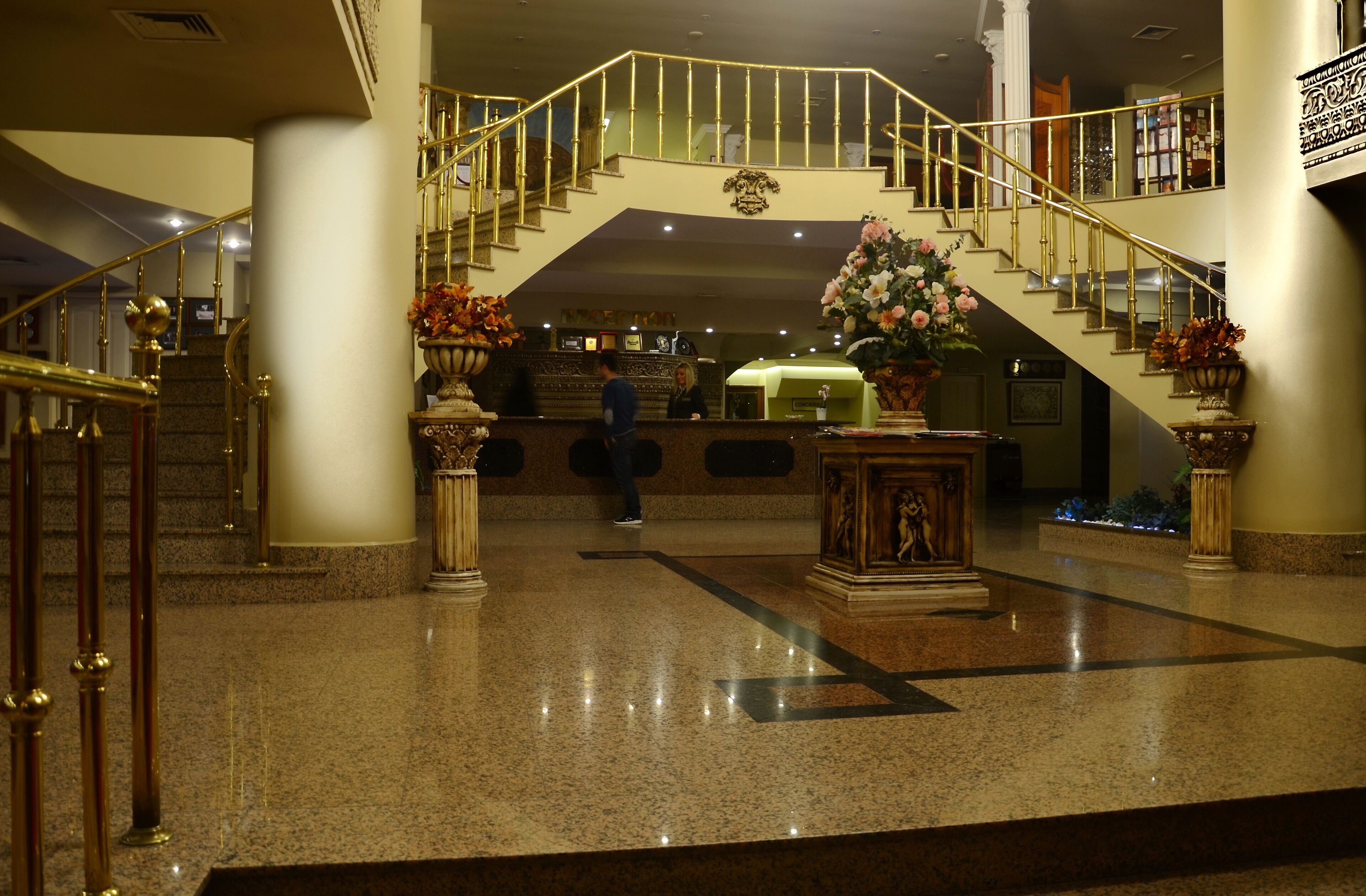 A grand lobby with a marble floor, a staircase with golden railings, and a reception desk.