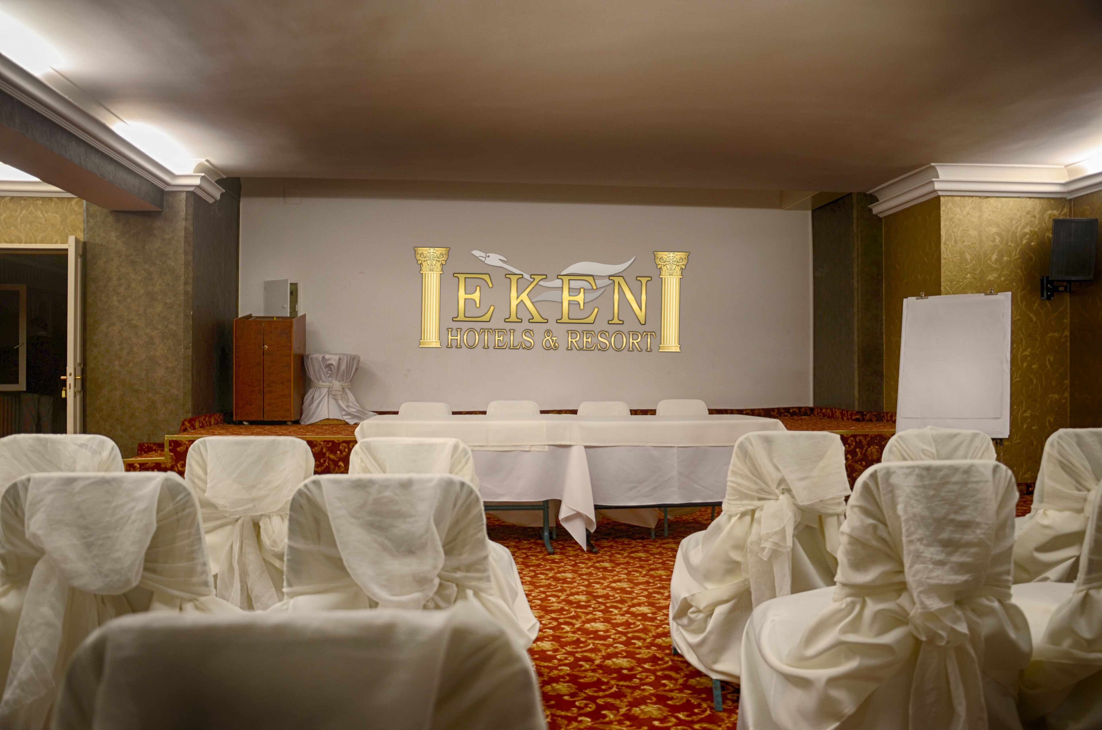 Un salón de conferencias con un escenario, sillas blancas con cintas y un cartel que dice 