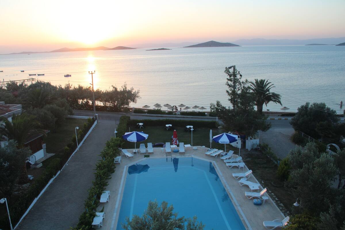 A swimming pool with lounge chairs and umbrellas, surrounded by greenery, with a view of the sea and distant islands at sunset.