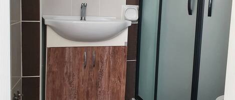 Un baño con lavamanos blanco, tocador de madera y cabina de ducha con mampara de vidrio.