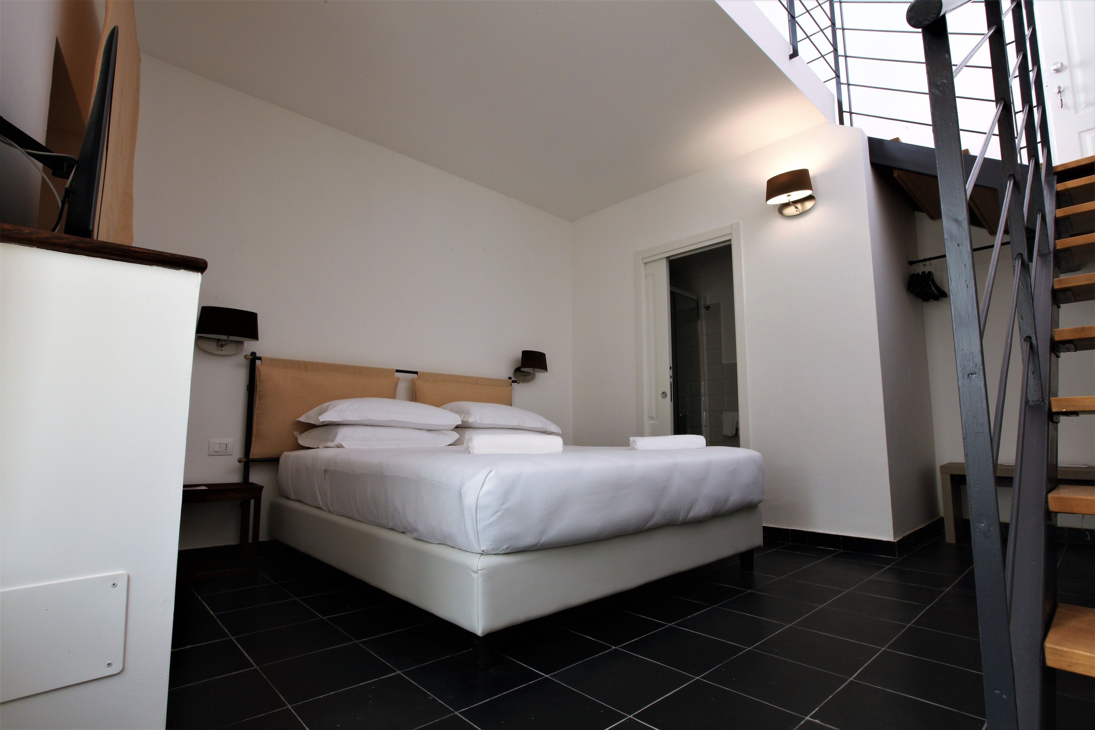 A modern bedroom with a large bed, a small bedside table, a wall-mounted lamp, and a staircase leading to an upper level.