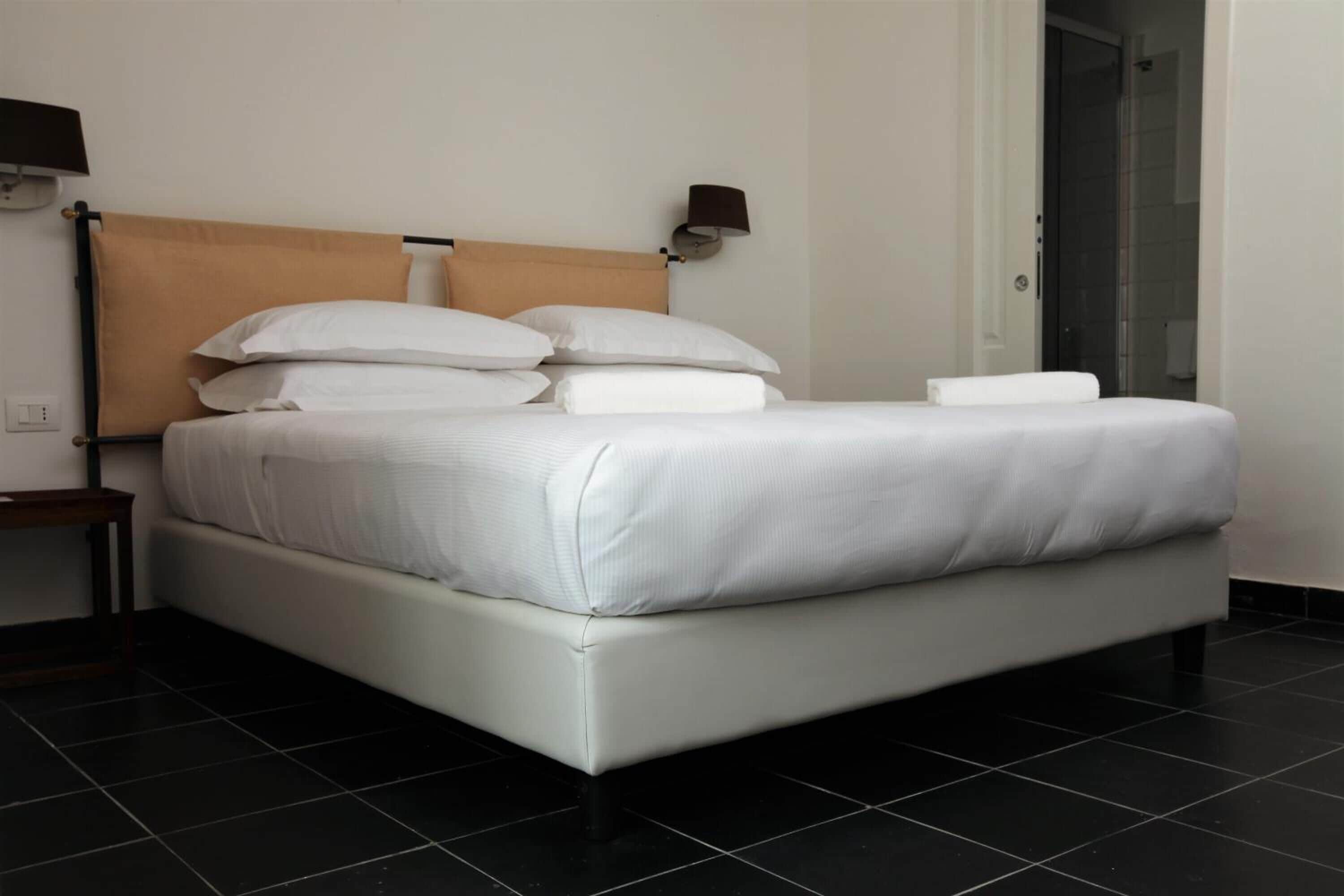 Una cama bien hecha con sábanas blancas y un cabecero, una mesita de noche con lámpara, y un piso de baldosas.
