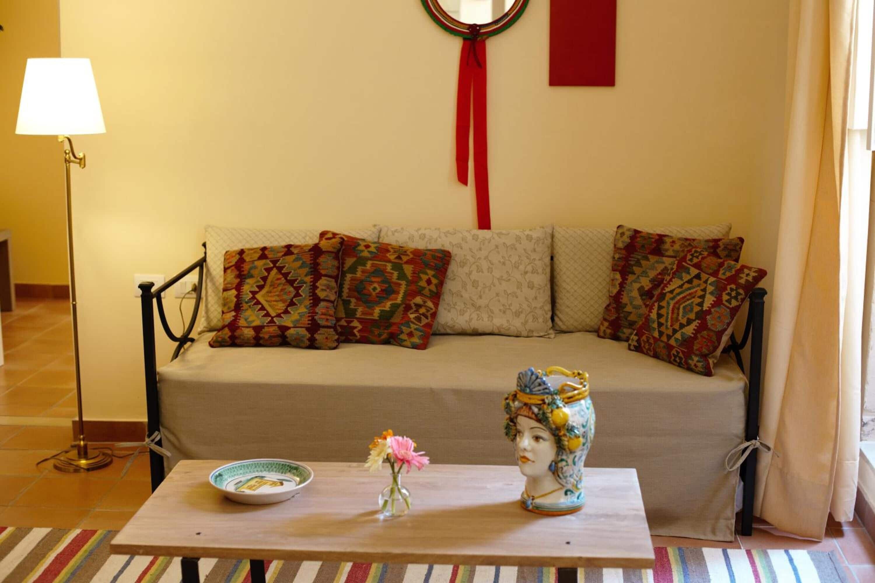 Una sala de estar con un sofá, cojines estampados, una mesita con objetos decorativos y una lámpara de pie.