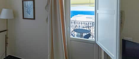 A room with a door leading to a balcony with a view of the sea, a framed picture on the wall, and a lamp.