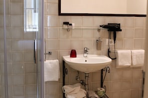 Un baño con cabina de ducha de vidrio, un lavamanos blanco con dispensador de jabón rojo y dos toallas blancas colgadas de ganchos.