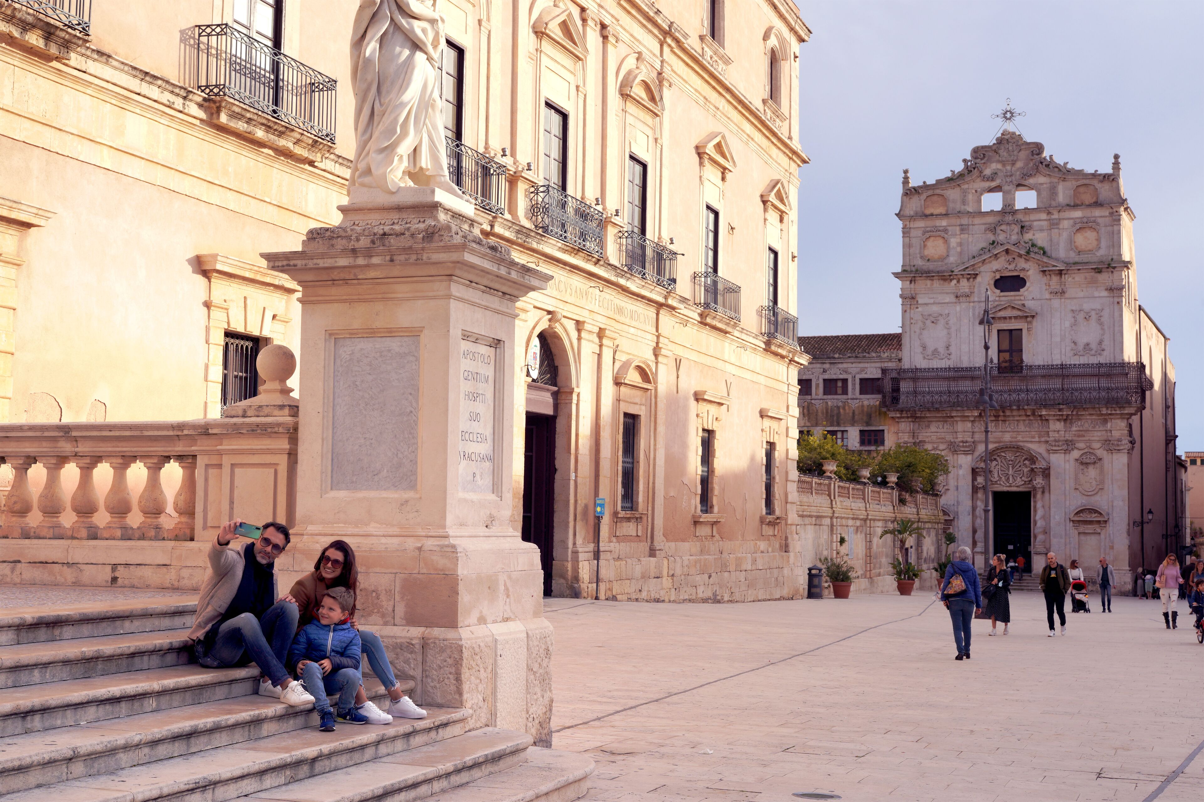 A historic building with a statue in front, a large church in the background, and people walking around.