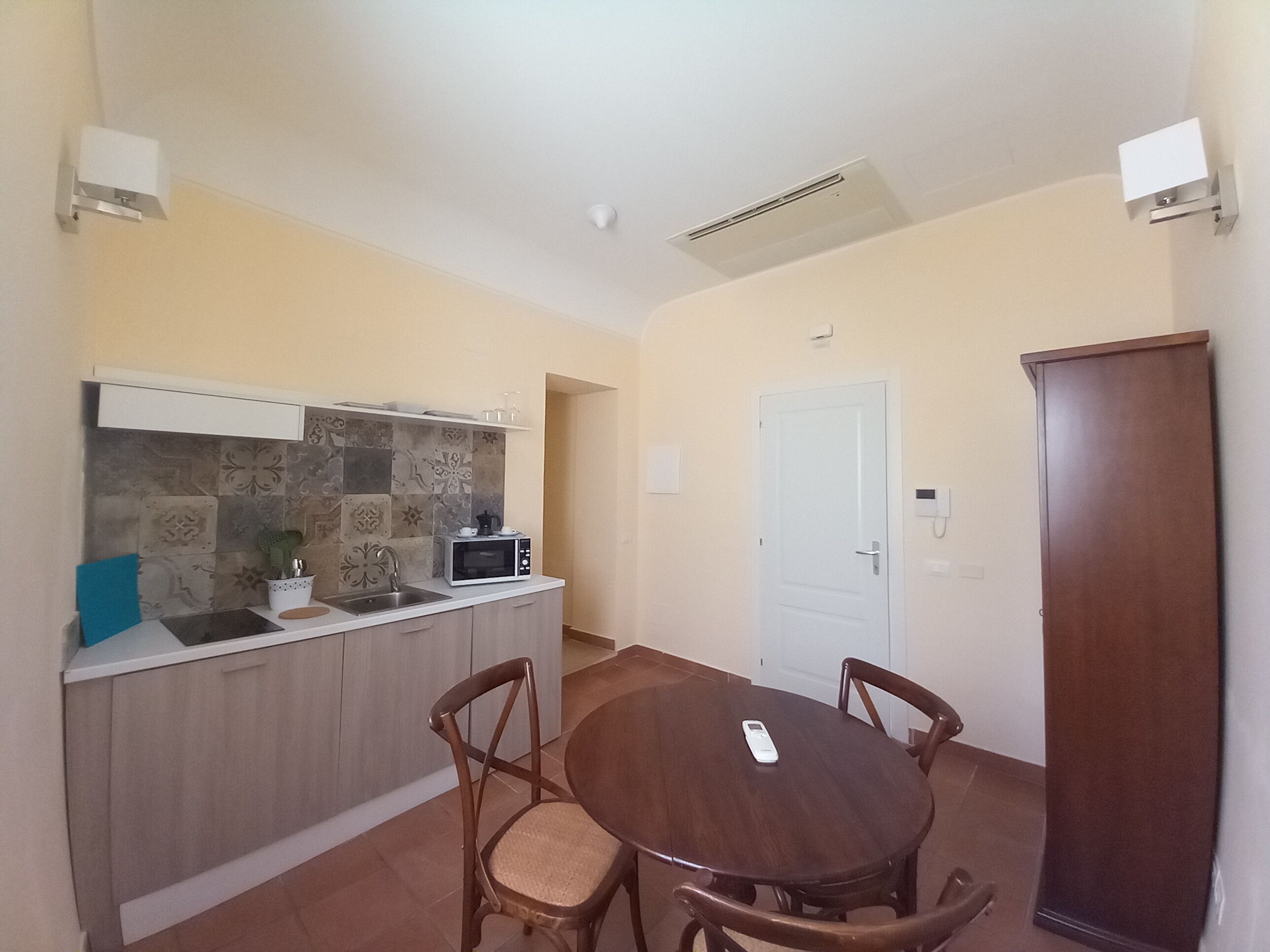 A compact kitchen with a microwave, sink, and decorative tiles. There is a dining table with chairs and a door in the background.