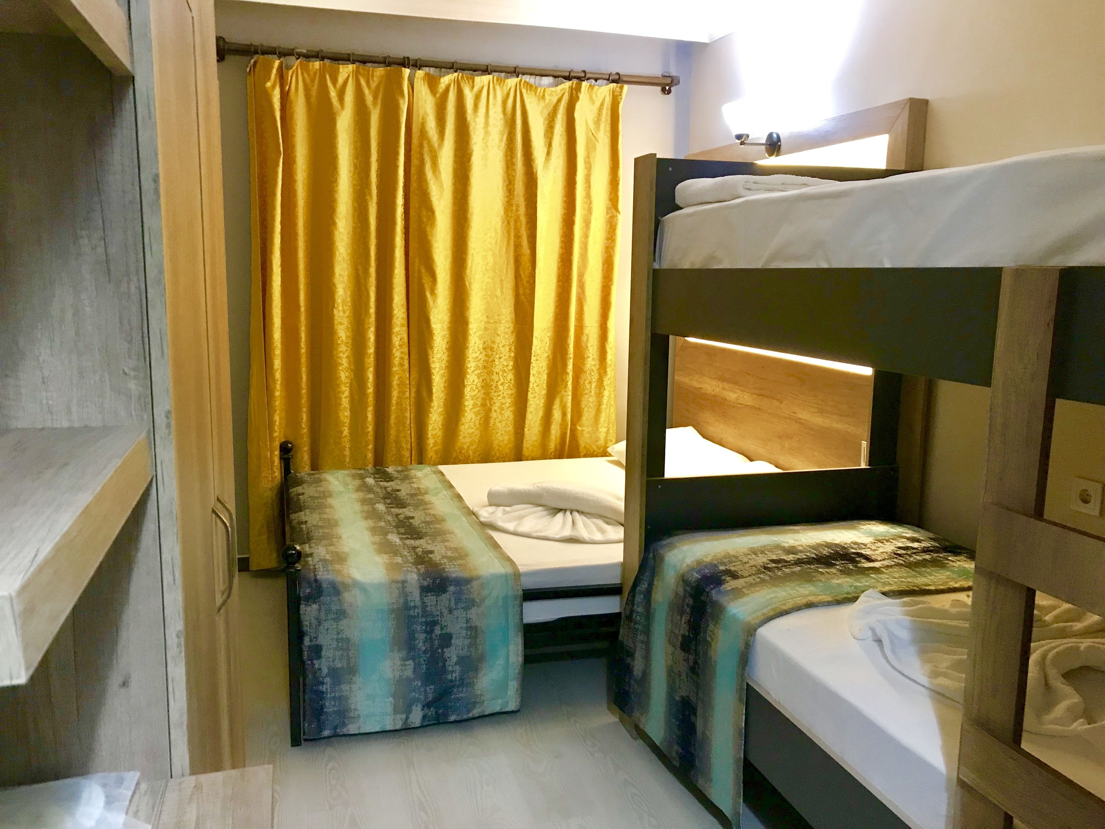 Una habitación pequeña y funcional con dos literas, una cama individual, una ventana con cortinas amarillas y un cabecero de madera.