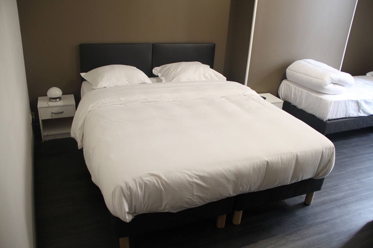 Een hotelkamer met een groot bed, een nachtkastje met een lamp, en een kleiner bed in de hoek.