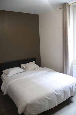 Un lit bien fait, avec du linge de lit blanc, dans une pièce aux murs sobres et dotée d’une fenêtre avec des rideaux.
