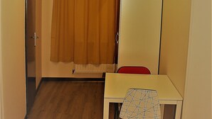 Une petite pièce avec un plancher en bois, une table blanche avec une chaise rouge, un meuble blanc et un rideau marron.