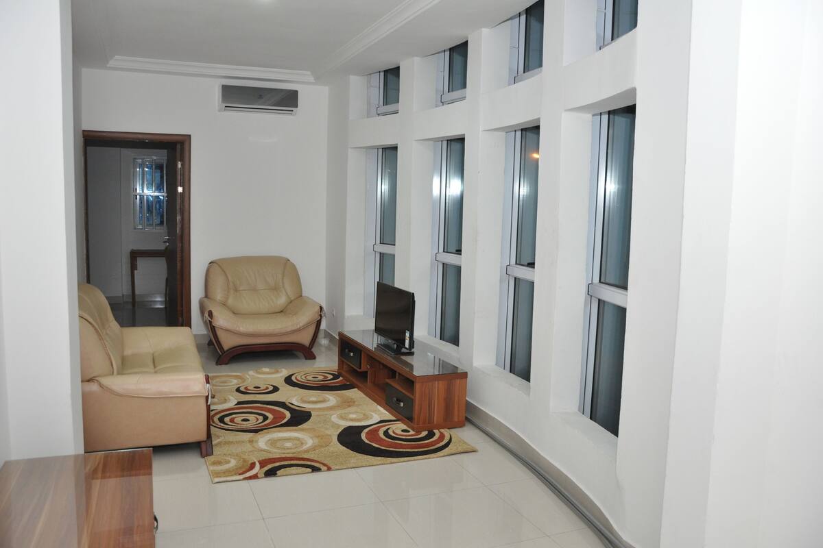 A living room with a beige sofa, a leather chair, a TV on a wooden stand, and a patterned rug.