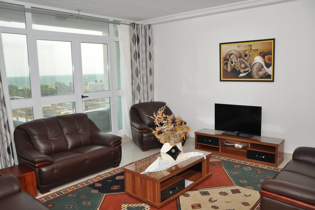 A living room with a brown leather sofa, a brown leather armchair, a glass coffee table with a vase, a flat-screen TV, and a patterned rug.