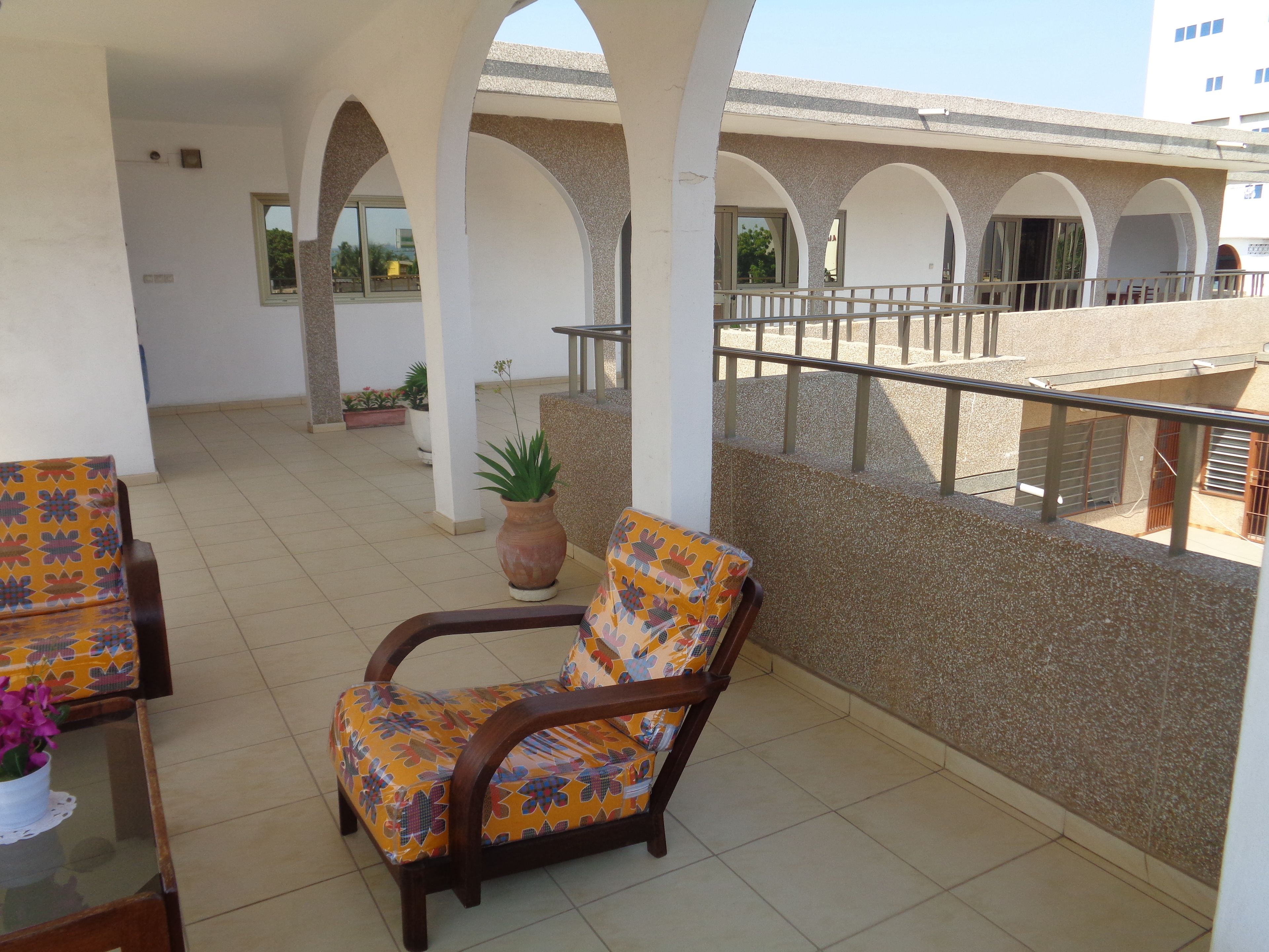 A covered outdoor seating area with a chair and table, a potted plant, and a view of a building with multiple arches.