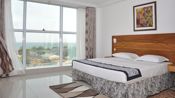 Une chambre avec une grande fenêtre, un lit recouvert de draps blancs, une tête de lit en bois et un tableau accroché au mur.