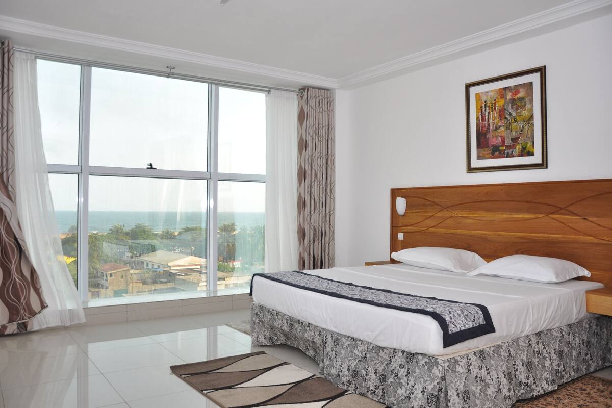 A bedroom with a large window, a bed with white bedding, a wooden headboard, and a painting on the wall.