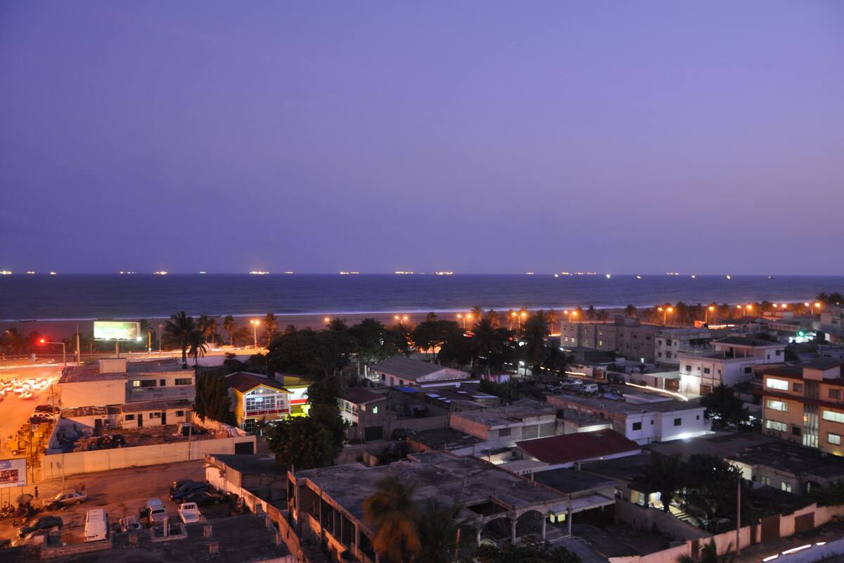 A coastal cityscape at dusk with buildings, a road, and a distant shoreline.