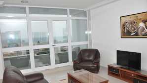 Un salon avec une grande fenêtre, un fauteuil en cuir, une table basse en bois, un téléviseur à écran plat posé sur un meuble en bois, et un tableau accroché au mur.