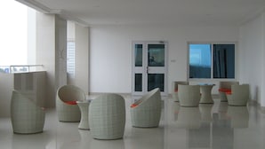 Une pièce spacieuse et moderne, dotée d’un plancher ciré, de plusieurs fauteuils en osier avec des coussins orange, et d’une grande fenêtre offrant une vue sur le ciel.