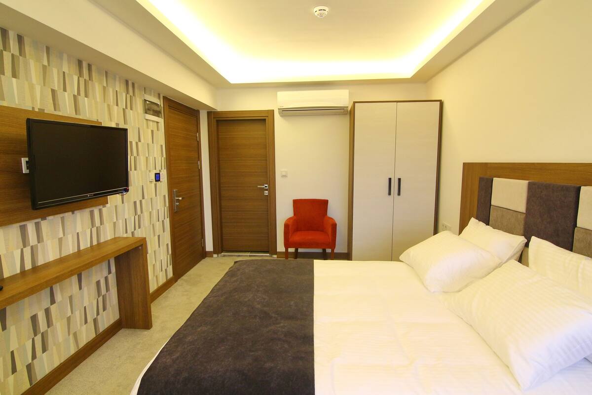 Una habitación de hotel con cama, televisión montada en la pared, una silla roja y un armario.