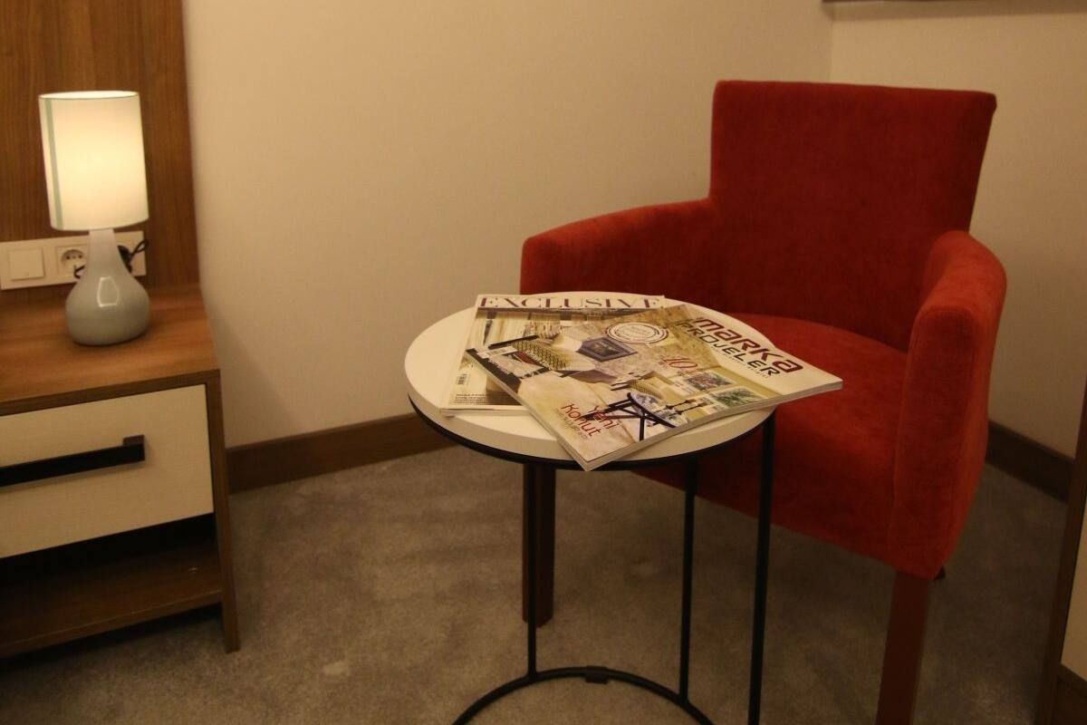 Habitación de hotel con un sillón rojo, una mesita redonda con revistas, una lámpara sobre una mesita auxiliar de madera y un cuadro en la pared.