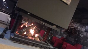 A fireplace with logs burning, surrounded by red chairs and a stone wall.