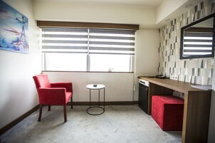 Una habitación pequeña y bien iluminada con una sola silla roja, una mesita redonda, un escritorio de madera y un reposapiés rojo. Un ventanal con persianas horizontales deja entrar luz natural.