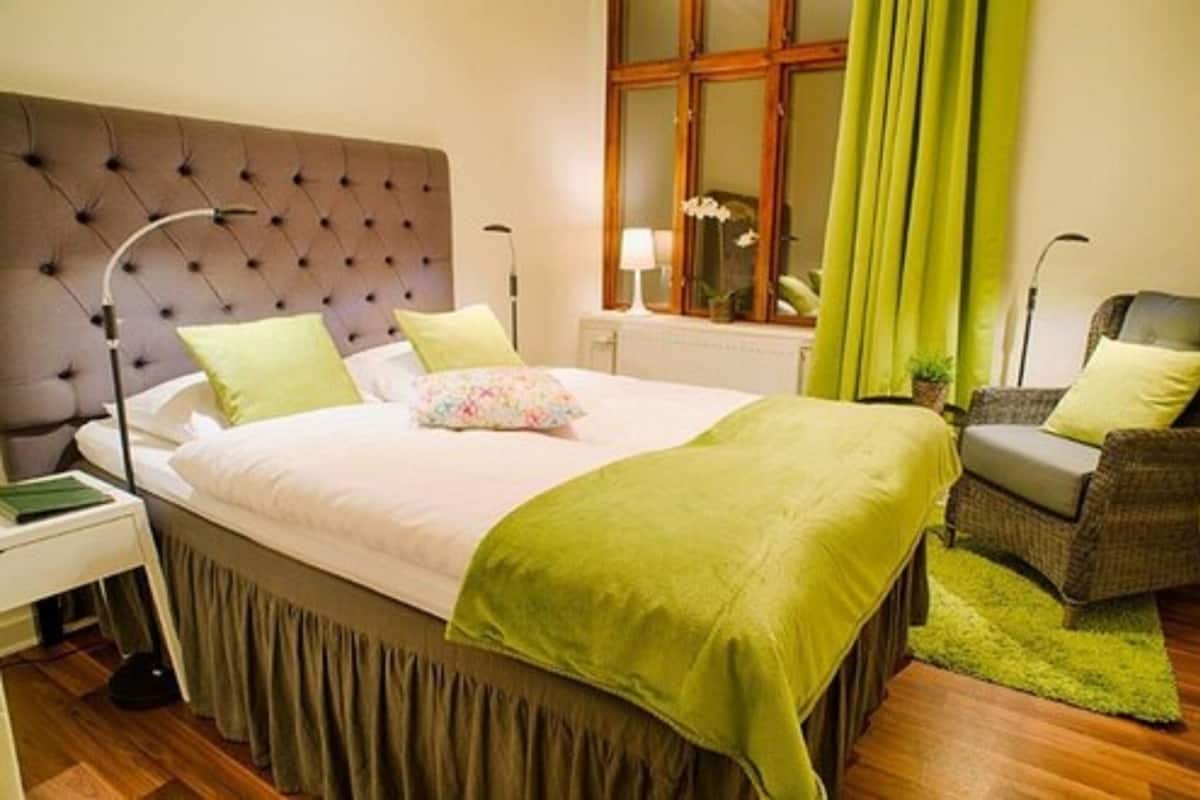 A neatly arranged bedroom with a tufted headboard, a bed with green and white bedding, a green throw blanket, a wicker chair, and a window with curtains.