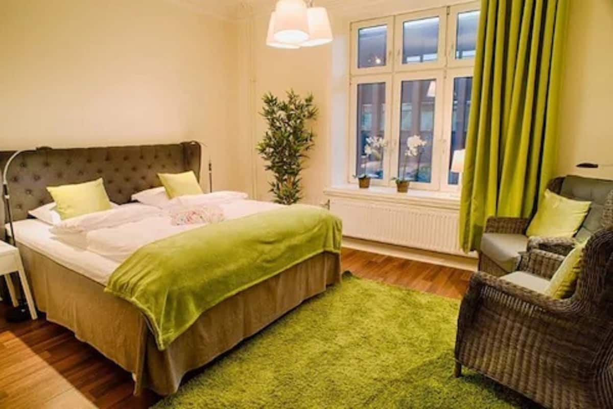 A bedroom with a bed, a green rug, a wicker chair, and a window with curtains.