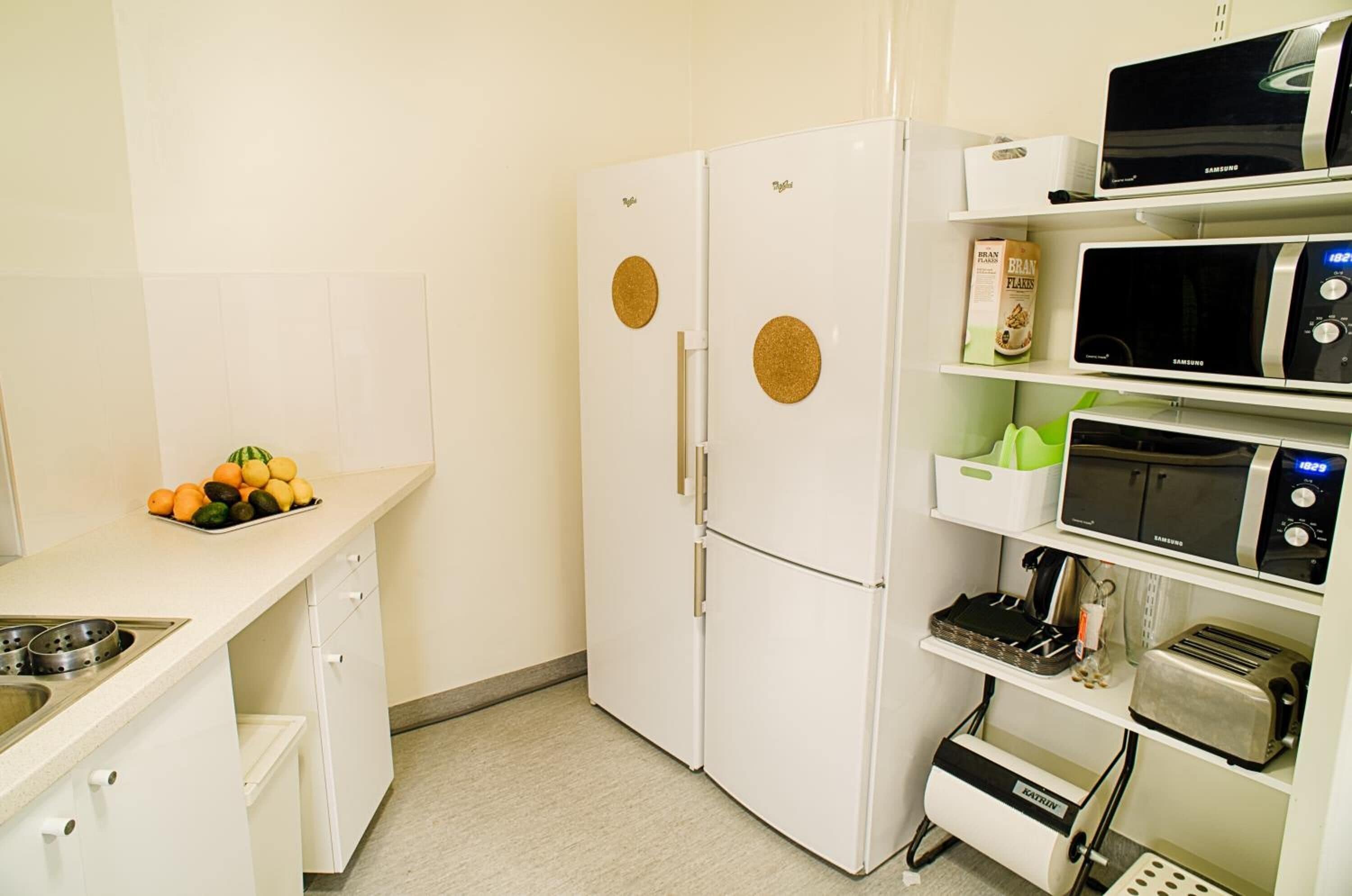 Een kleine keuken met een witte koelkast, magnetron en broodrooster. Op het aanrecht staat een fruitschaal en op de plank een pak havervlokken.
