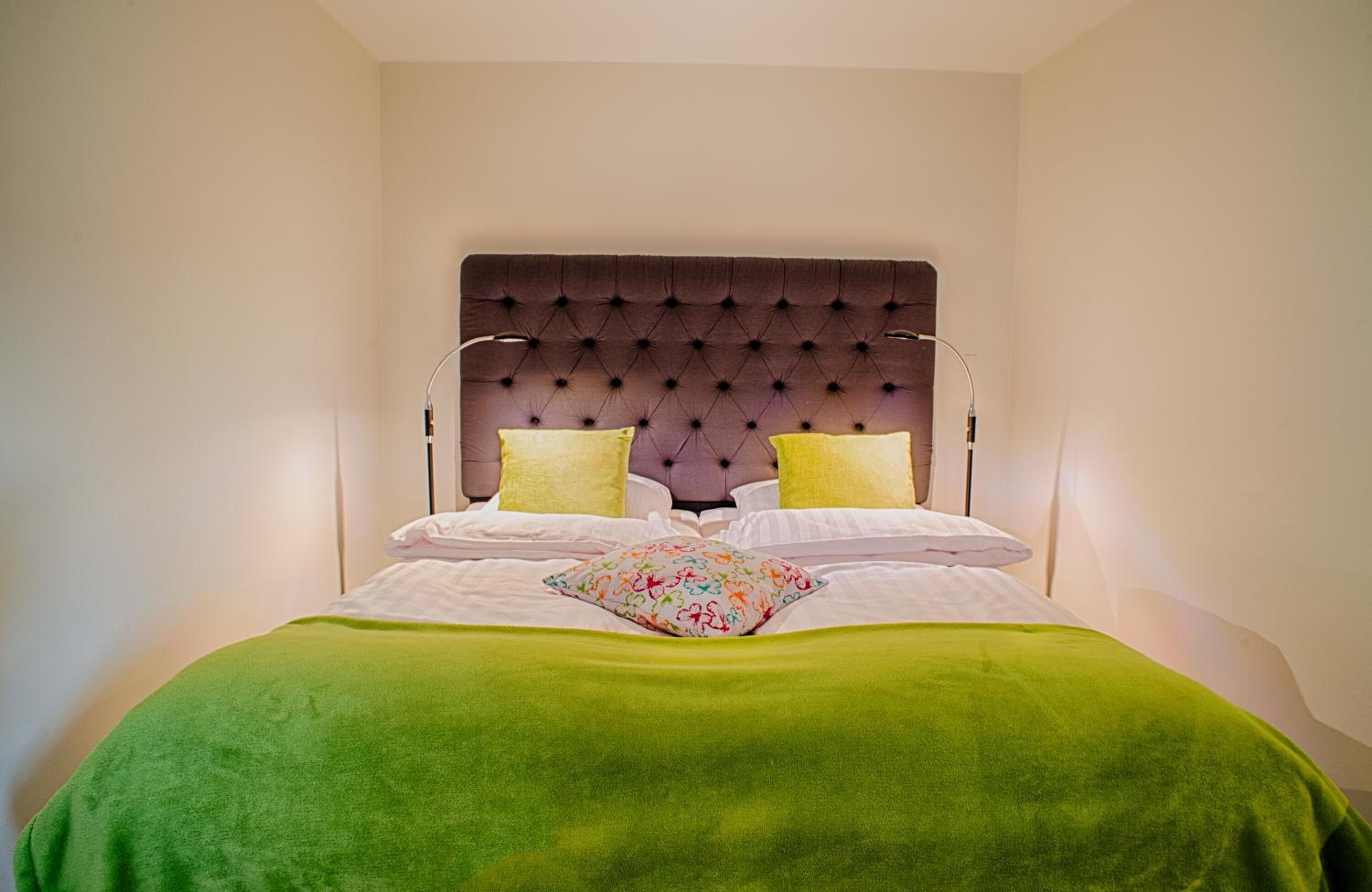 A neatly made bed with a green blanket and two pillows, a tufted headboard, and bedside lamps.