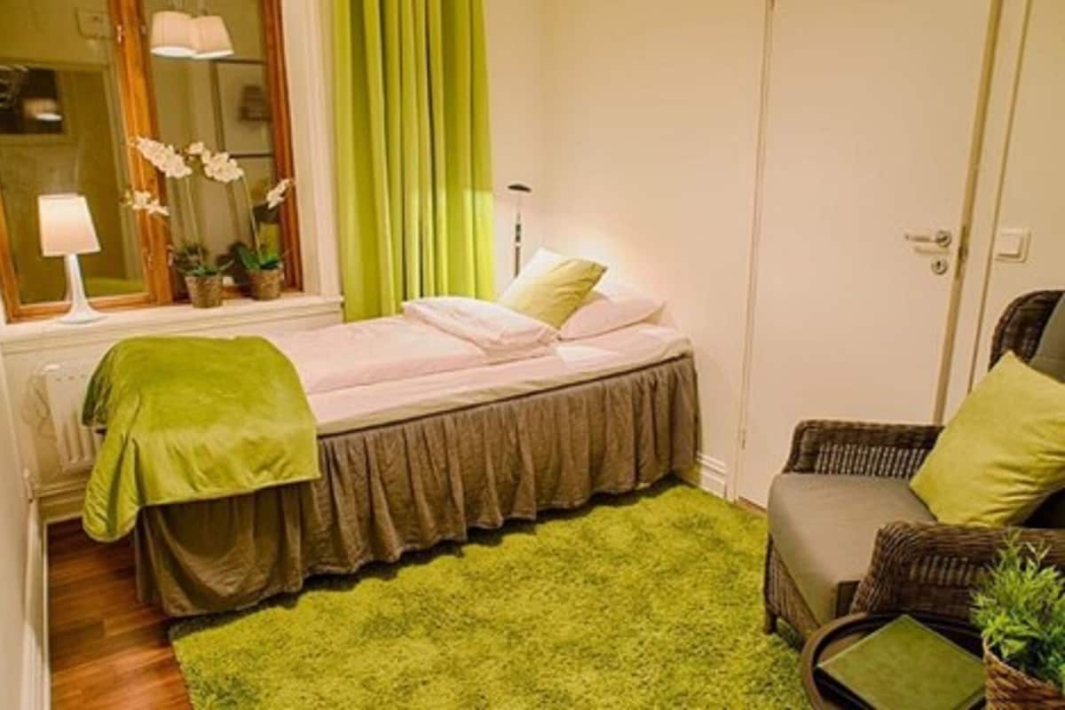 A small, cozy bedroom with a bed, a green rug, a chair, and a window with curtains.