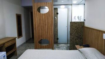A hotel room with a bed, a wooden wardrobe, a mirror, a desk, and a bathroom visible through an open door.