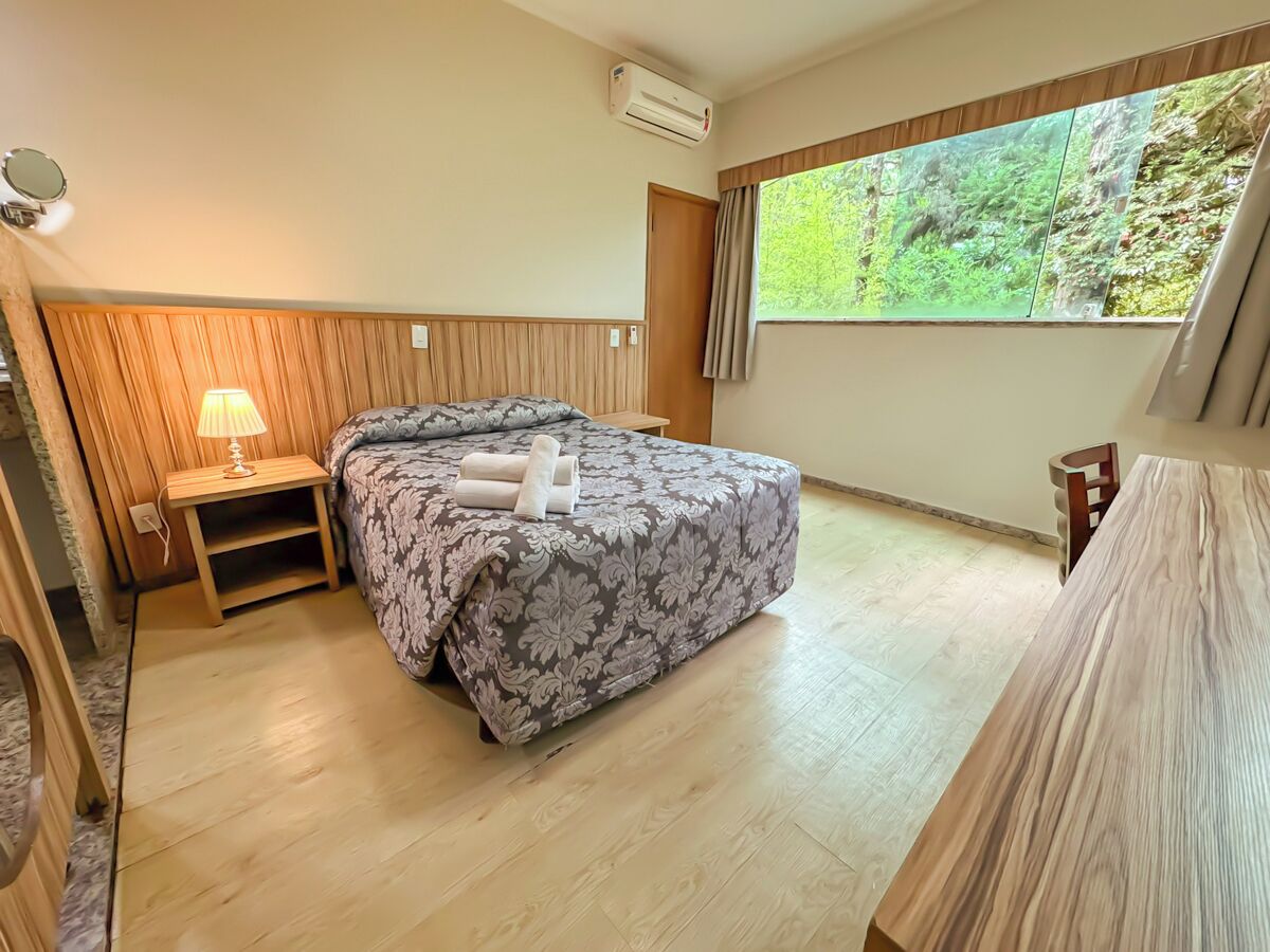 Quarto com cama, mesa de cabeceira com abajur, janela com cortinas e piso de madeira.